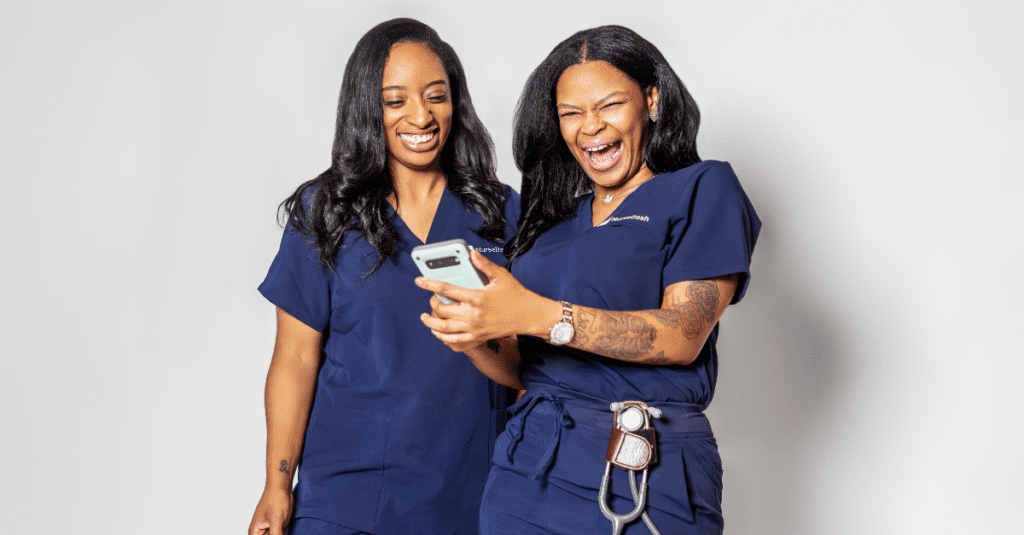 Two women nurses in navy blue scrubs smiling and holding cell phone in hand